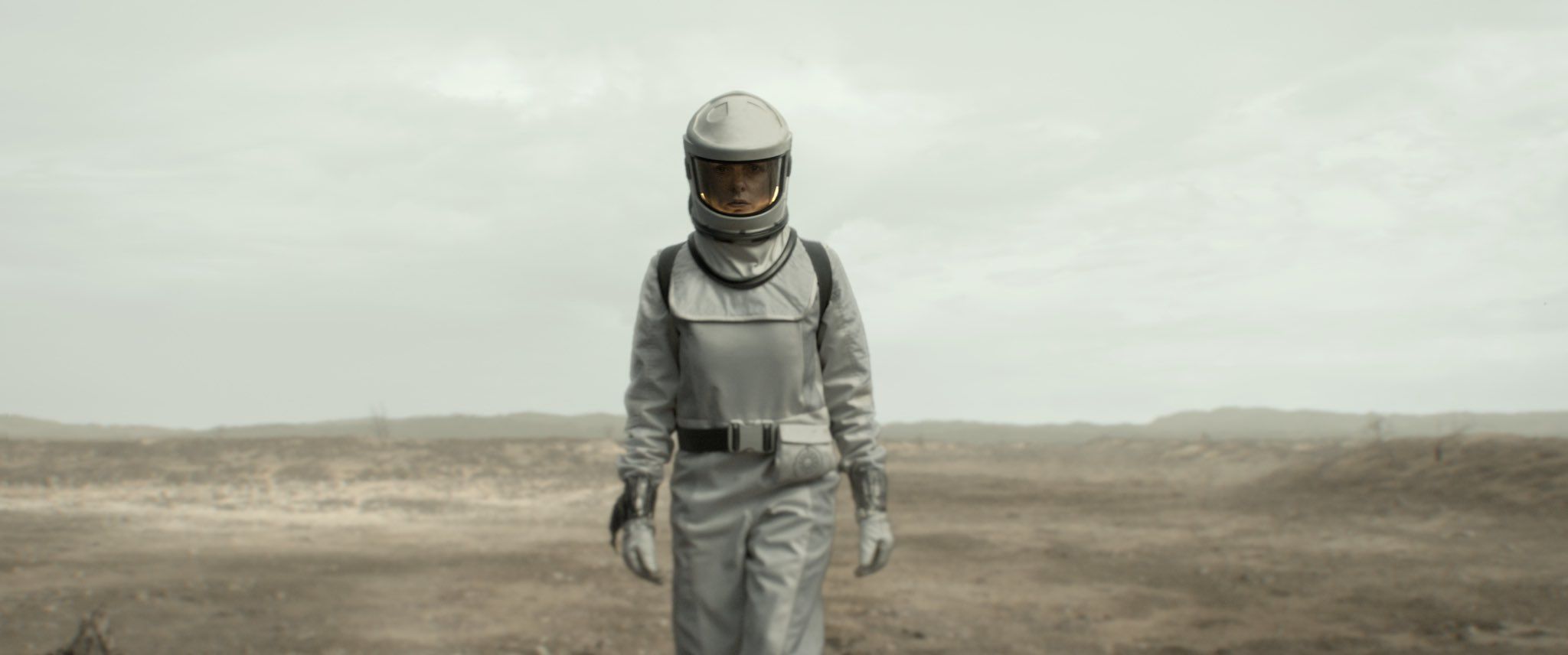 From Silo. A wide shot of Rebecca Ferguson as Juliette Nichols wearing a white environmental suit, standing in a desolate area. The ground behind her is barren, and the sky is empty. She has her hands at her sides and walks forward tentatively.